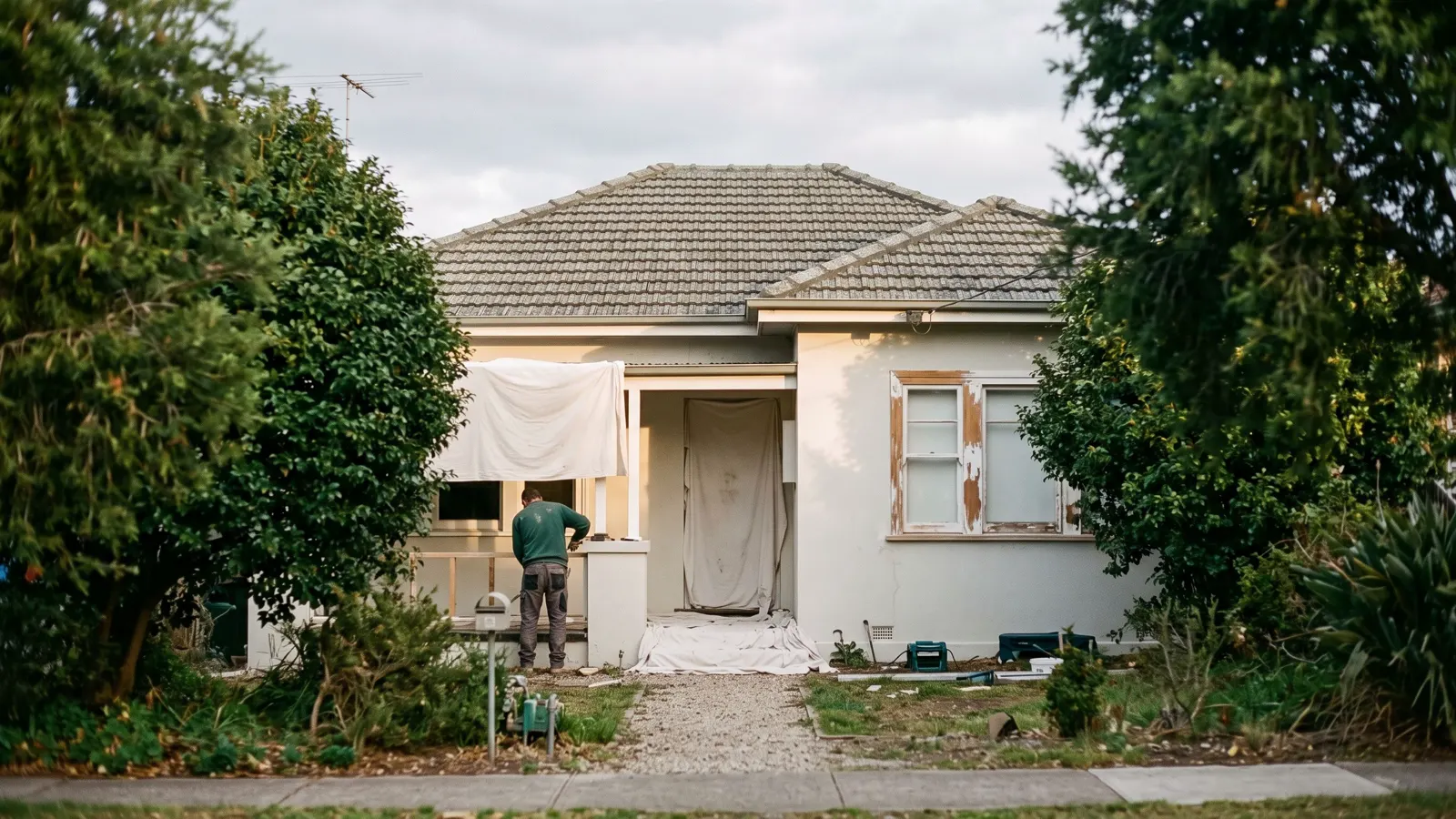 A settled suburban house under grown-in trees in Marlboro, NJ, dust sheets over the porch and a scraped window frame.